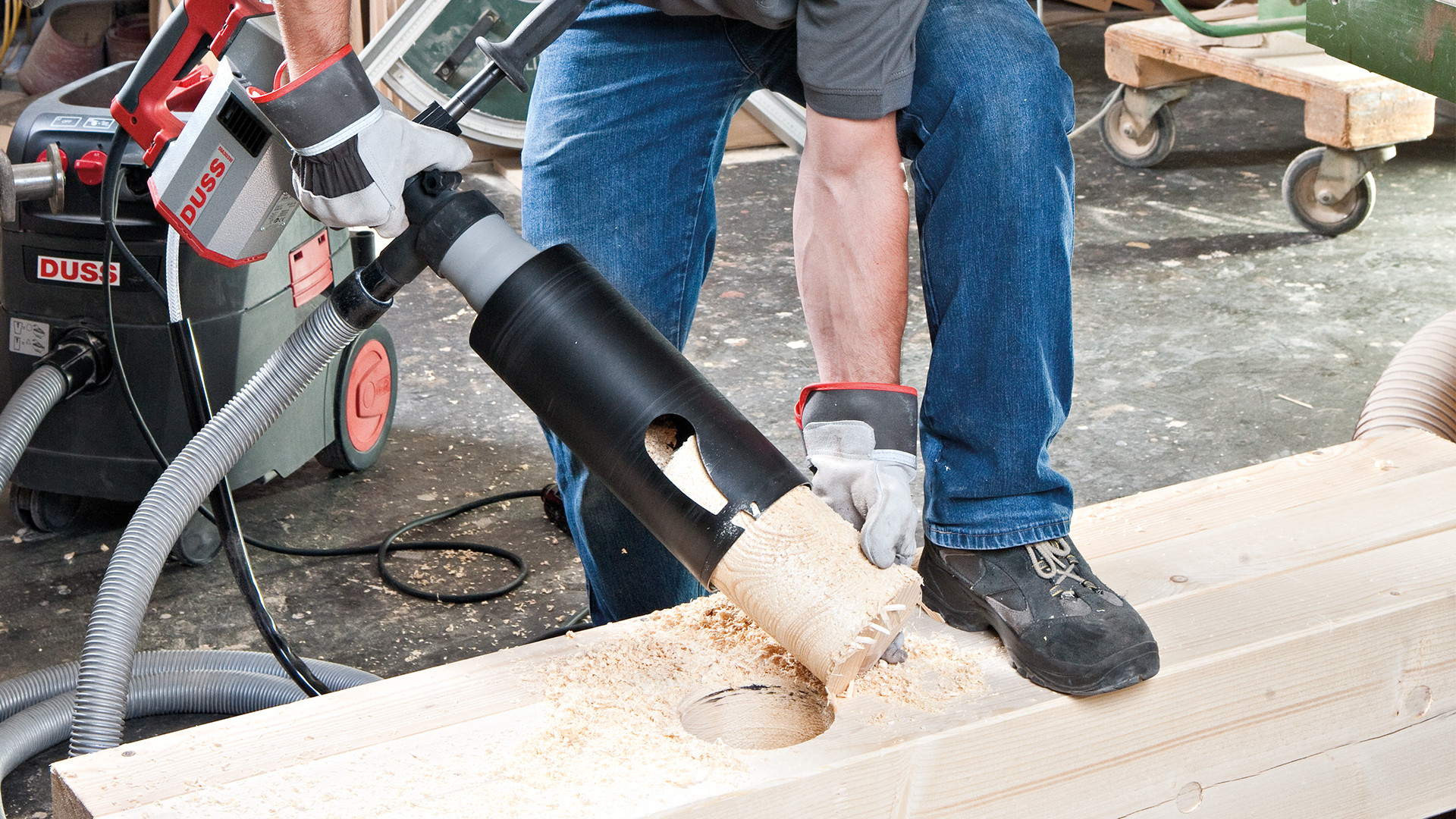 Professional woodworking machine drilling a large hole in a timber beam using a wood cutter tool, producing wood chips during structural timber processing